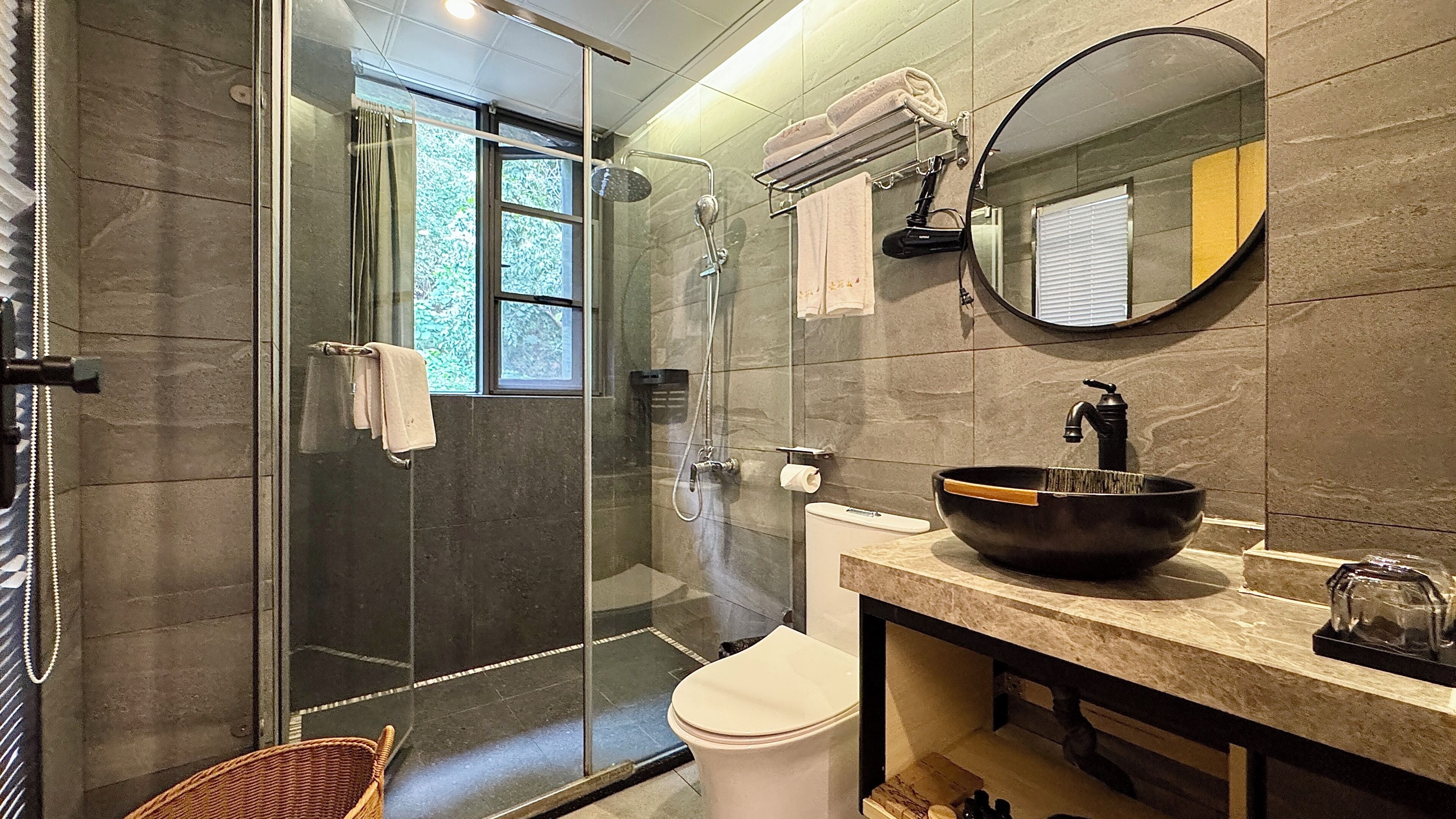 A modern bathroom with a glass-enclosed shower, a round mirror, a black vessel sink, and a toilet.