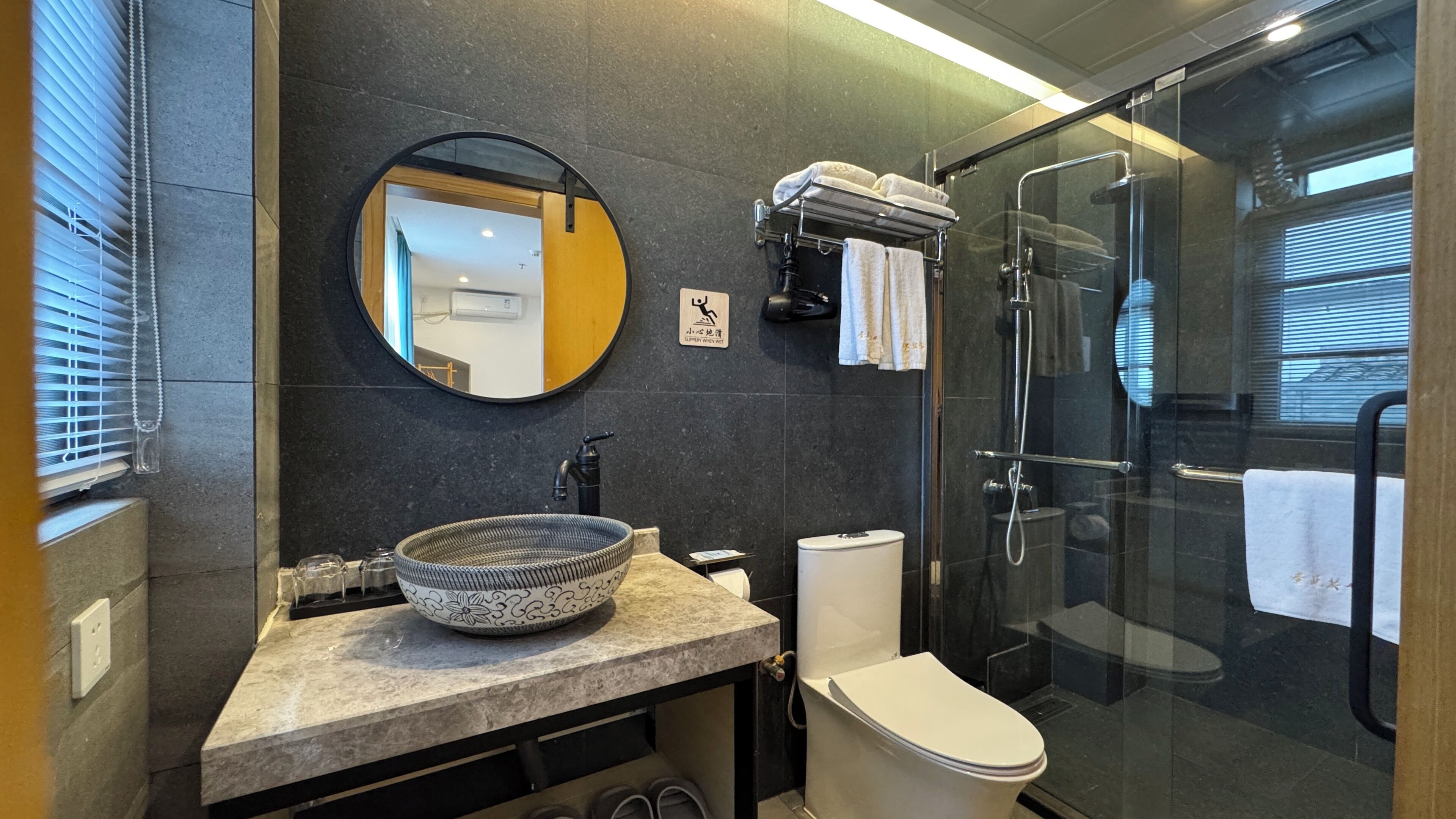 A bathroom with a round mirror, a sink with a decorative bowl, a toilet, and a glass-enclosed shower area.
