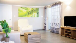 Un salon moderne avec un canapé blanc, une table basse en bois, un téléviseur à écran plat posé sur un meuble en bois, une grande peinture abstraite et une plante en pot.