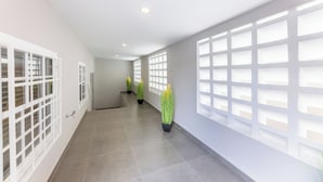Un couloir doté d’un sol carrelé, de murs blancs et de grandes fenêtres équipées de stores blancs.