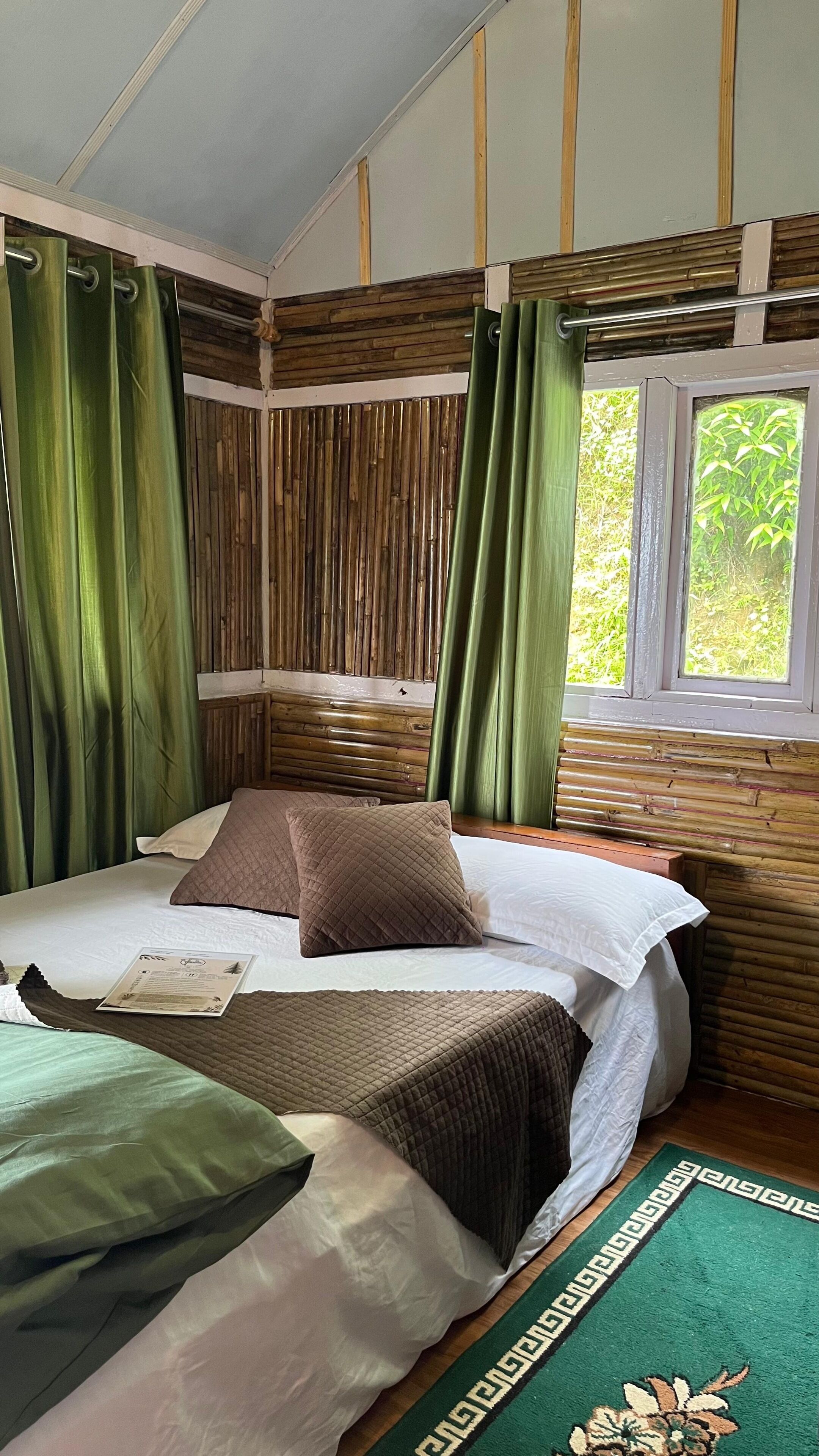A room with a bed, green curtains, a window with a view of greenery, and a wooden wall.