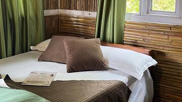 A room with a bed, green curtains, a window with a view of greenery, and a wooden wall.