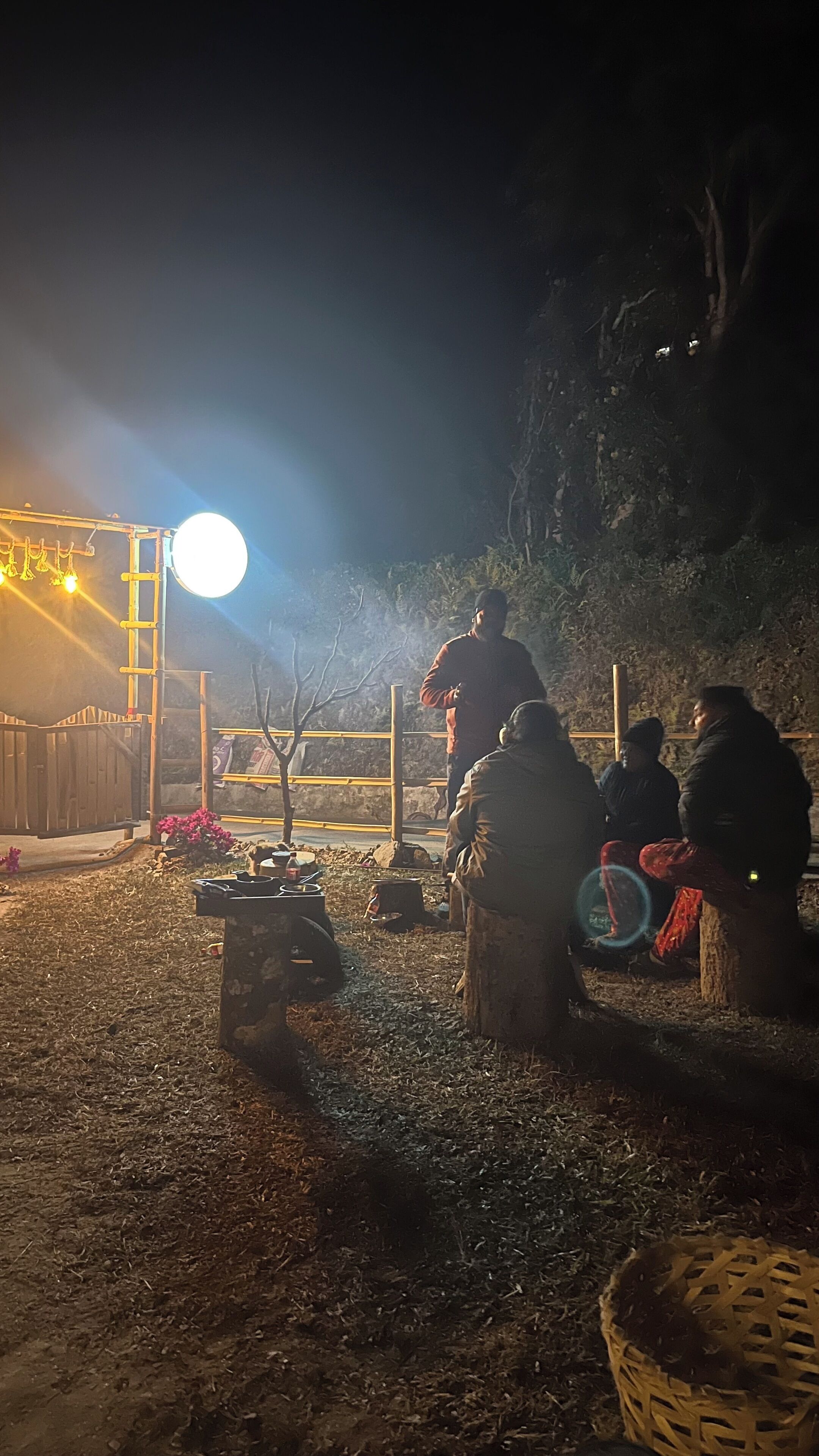 A night scene with a group of people gathered around a campfire, a wooden structure with a ladder, and a large light source in the background.