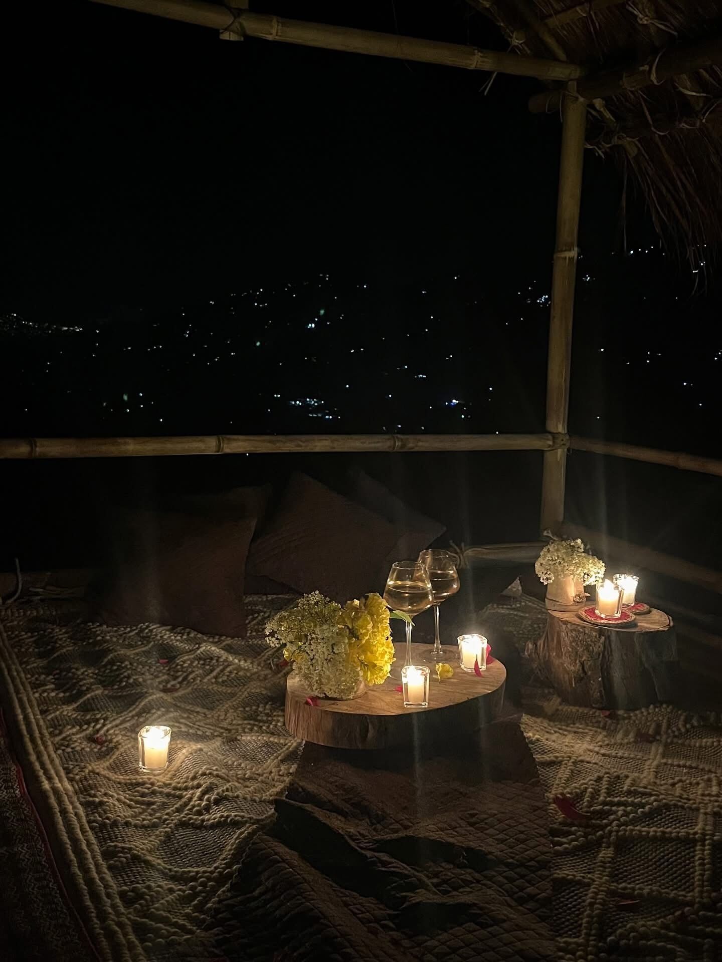 A cozy outdoor seating area with a table set for two, featuring candles, glasses, and flowers.