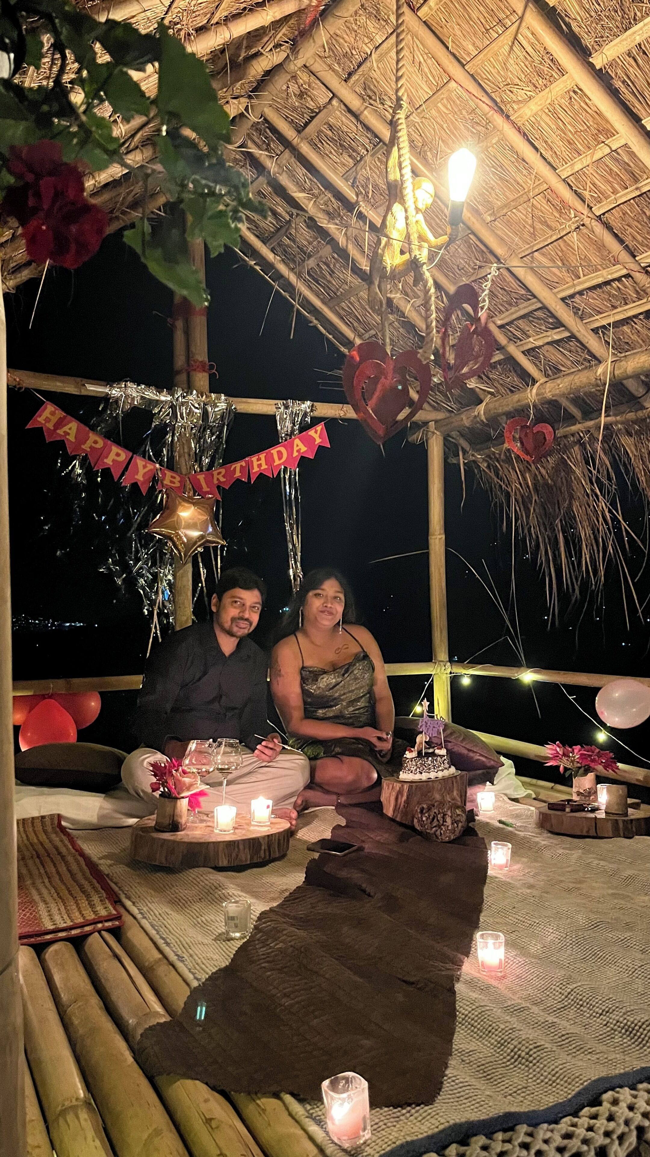 Un couple est assis sous une structure dont le toit est fait de chaume, devant une table dressée pour un repas, avec des bougies et un gâteau d’anniversaire.