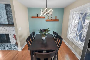 A dining area with a dark wooden table, six chairs, a potted plant, and two wooden shelves with decorative items. - Modern 3BR West Omaha, Private Pool now open, Fenced Yard, W/D, Pet-Friendly (Omaha)