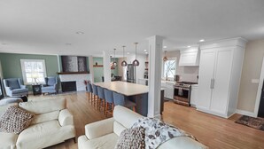 Uma sala de estar moderna com sofá e poltronas, área de jantar com mesa e cadeiras, e cozinha com armários brancos e eletrodomésticos de aço inoxidável.