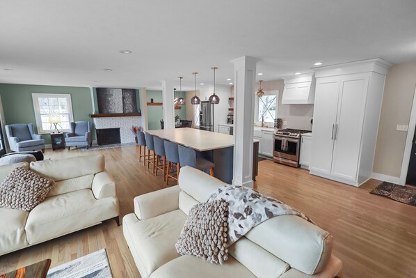 A modern living room with a sofa and armchairs, a dining area with a table and chairs, and a kitchen with white cabinets and stainless steel appliances. - Modern 3BR West Omaha, Private Pool now open, Fenced Yard, W/D, Pet-Friendly (Omaha)