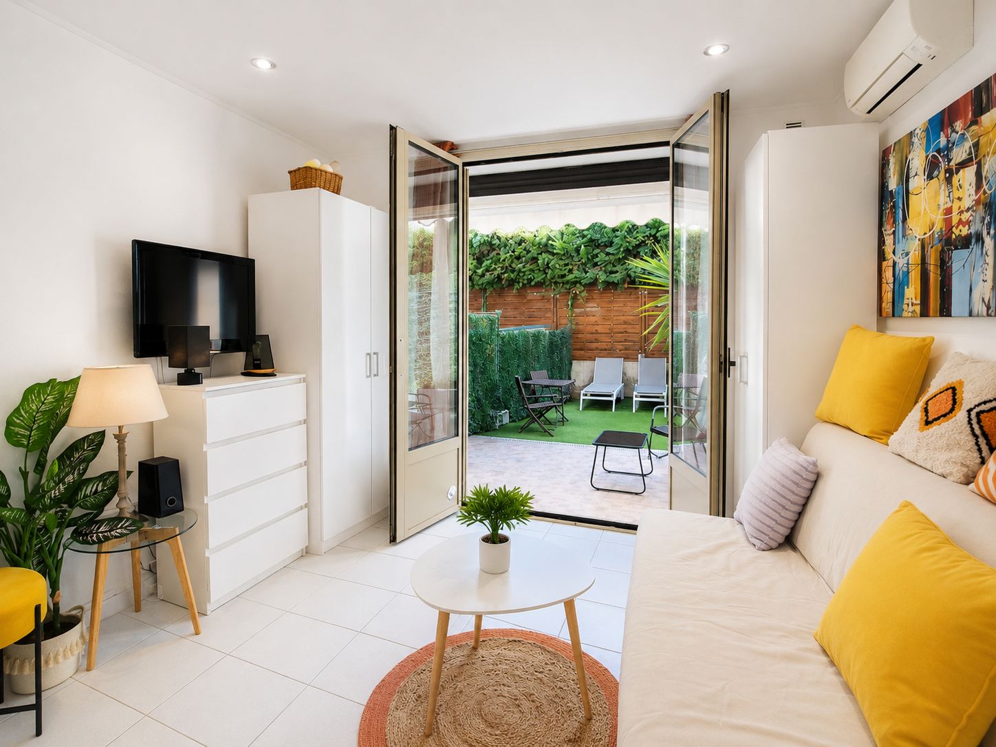 Una sala moderna con sofá, televisión y puerta de cristal que da a un patio con mobiliario exterior.