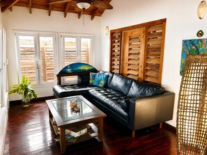 A living room with a leather sofa, a glass coffee table, wooden shutters, and a painting on the wall. - Dancing Oaks 3BR Tropical Retreat · Saltwater Pool · Private Dock (Punta Blanca)