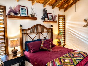 A bedroom with a wooden headboard, a red bedspread, and a colorful quilt. There are two bedside tables with lamps, a shelf with decorative items, and a window with blinds.