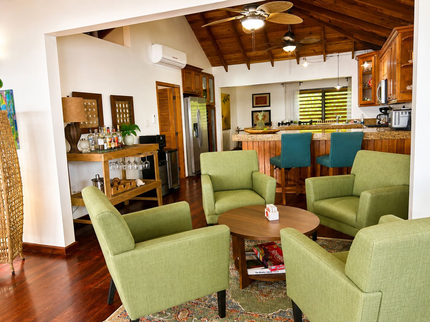 A spacious living area with a wooden ceiling, a dining table with four green chairs, a kitchen with wooden cabinets, and a bar area with two blue chairs.