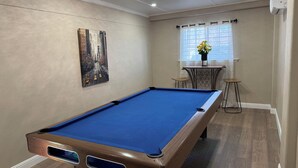 Een pooltafel in een kamer met een stadsgezichtschilderij, een klein tafeltje met een bloempot en een raam met gordijnen.
