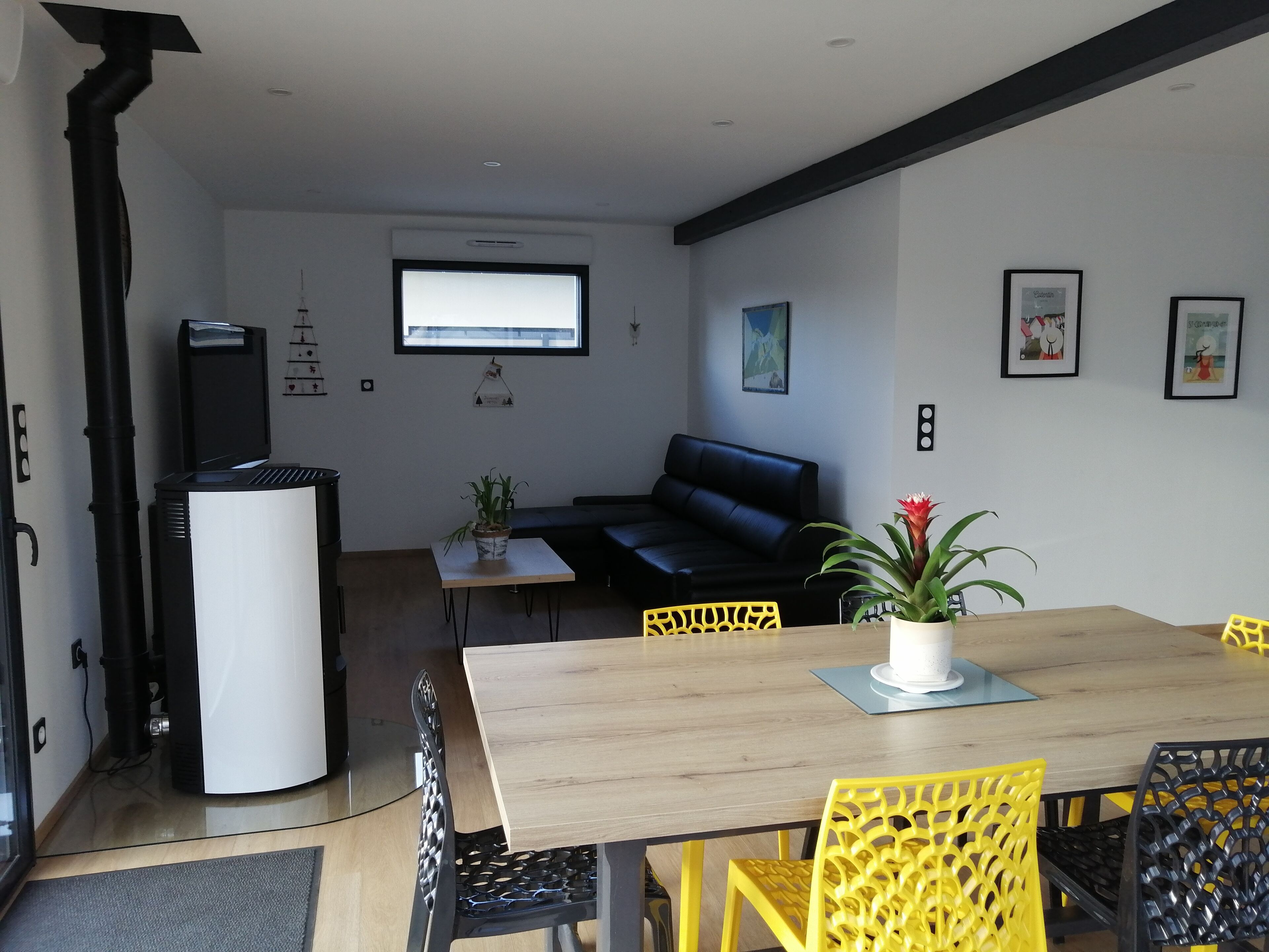 Uma sala de estar moderna com um sofá de couro preto, uma mesa de jantar de madeira com cadeiras amarelas, uma geladeira branca e uma planta em vaso.