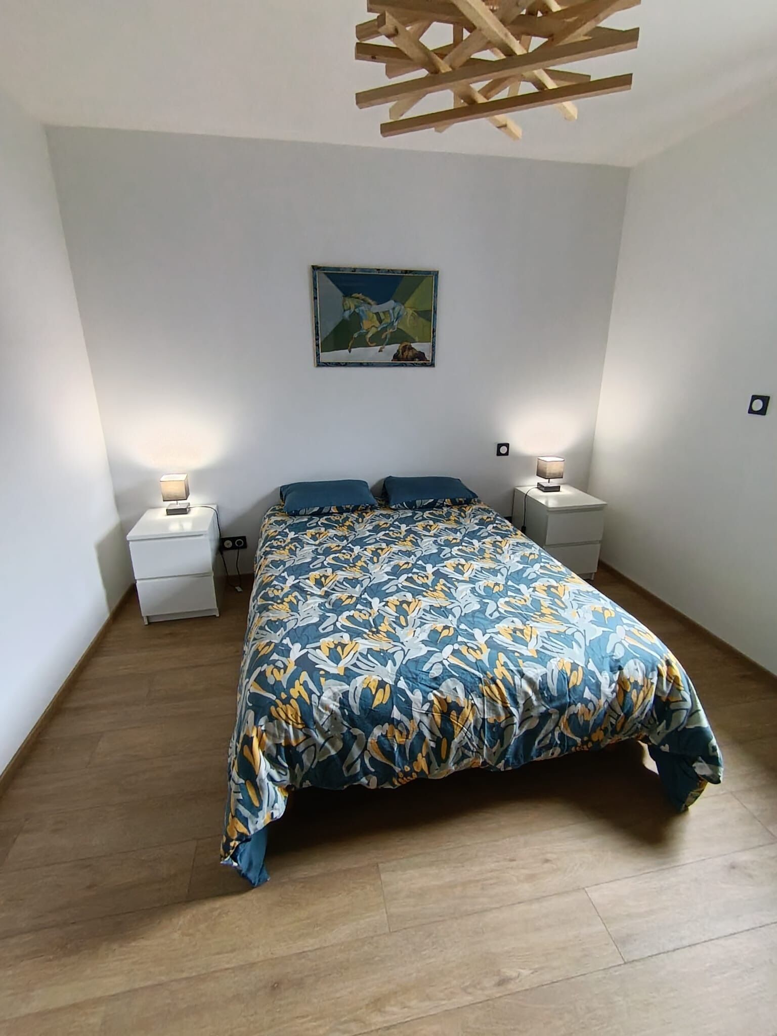 A bedroom with a bed, two bedside tables, a wall-mounted painting, and a wooden ceiling light fixture.
