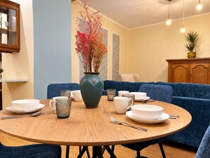 A round wooden table with white plates, cups, and cutlery, surrounded by a blue sofa and a vase with red flowers. - 1-bedroom in Old Town (Tallinn)