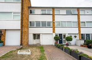 A two-story residential building with white exterior walls, a brick section, and a small front garden with potted plants and a flower bed. - Beautiful Orpington Home w Pool Table (Orpington)