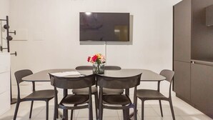 Ein modernes Esszimmer mit einem runden Tisch, sechs Stühlen, einem wandmontierten Flachbildfernseher und einer Tischvase mit Blumen.