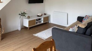 A modern living room with a wooden staircase, a flat-screen TV mounted on the wall, a grey sofa, and a white console table with decorative items. - Family home with spa bath in one of the most beautiful villages in France ! (Rochefort-en-Terre)