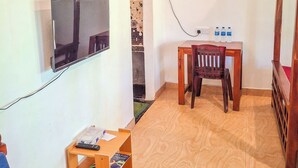 Una stanza con un televisore montato a muro, un tavolo in legno con una sedia, una piccola mensola con oggetti e una ventola da pavimento.