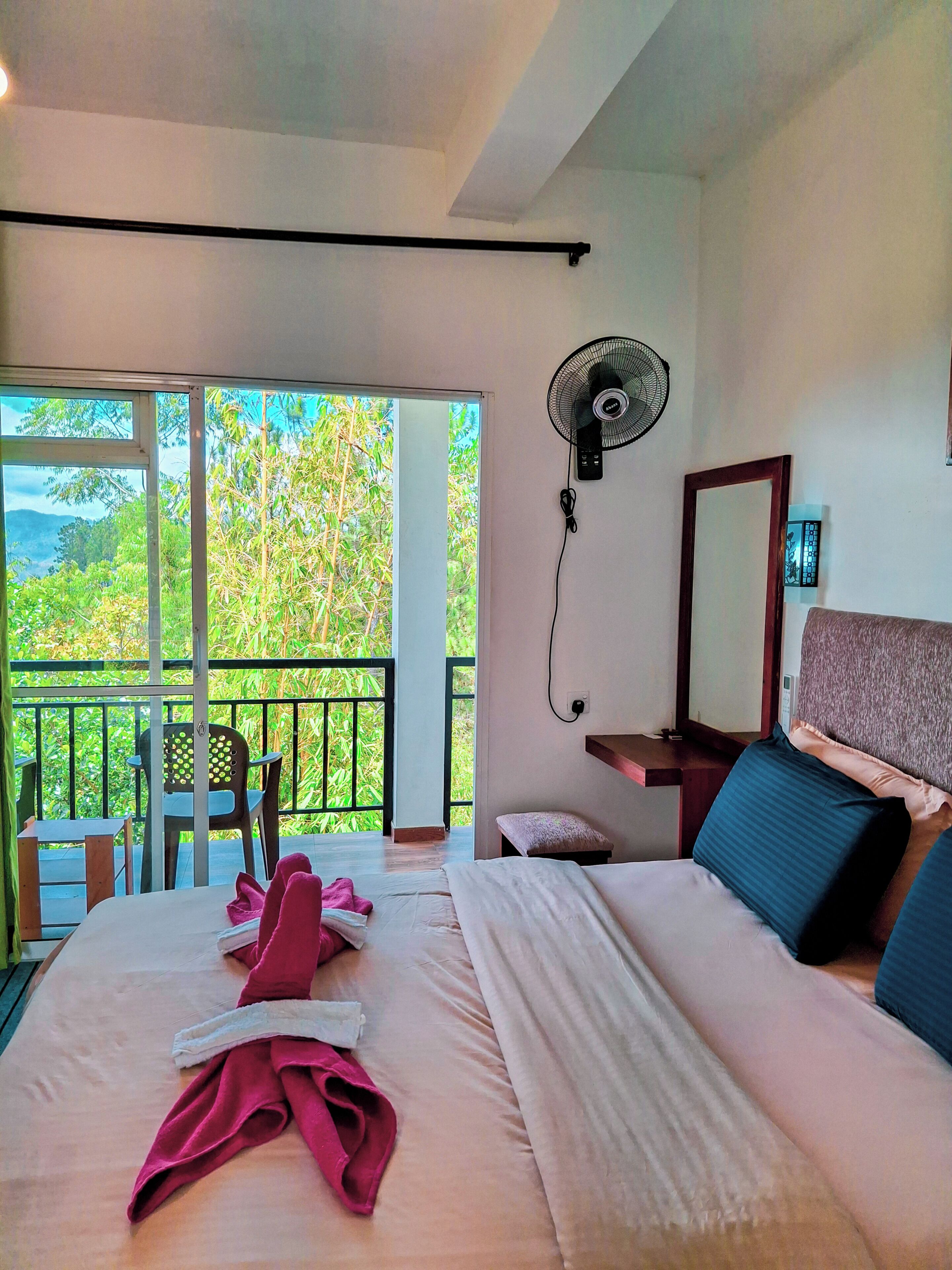 A hotel room with a bed, a desk, a chair, a balcony with a view of greenery, and a wall-mounted fan.
