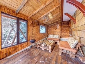 Interior acogedor de una cabaña con paredes y suelos de madera, un ventanal con vistas a árboles cubiertos de nieve, un banco de madera y una pequeña mesa.