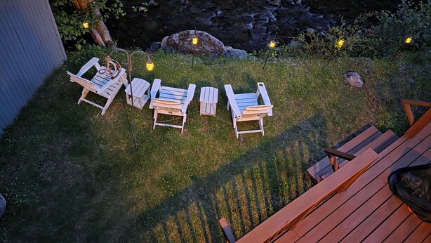 Una terraza con cuatro sillas Adirondack blancas y una mesa de madera, con vistas a un arroyo pedregoso y un espacio verde con luces pequeñas.
