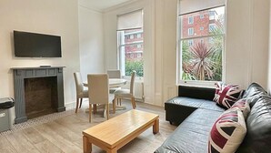 A modern living room with a fireplace, a sofa with British flags, and a view of a red brick building through large windows. - Bright Apartment Stockwell Station Nearby (London)