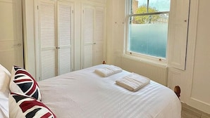 A bedroom with a large bed, white bedding, and a window with a view of buildings and trees. - Bright Apartment Stockwell Station Nearby (London)