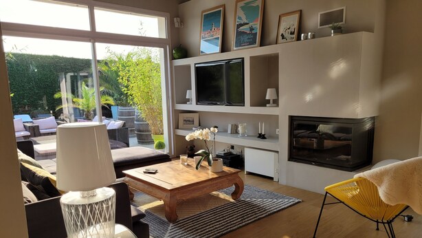 A modern living room with a sofa, coffee table, and TV. There is a fireplace and a large window with a view of greenery. - Magnificent contemporary Basque house with pisicine in a green setting (Ustaritz)