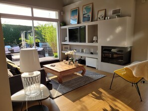 A modern living room with a sofa, coffee table, and TV. There is a fireplace and a large window with a view of greenery. - Stunning modern Basque house with a pool in a green setting   (Ustaritz)