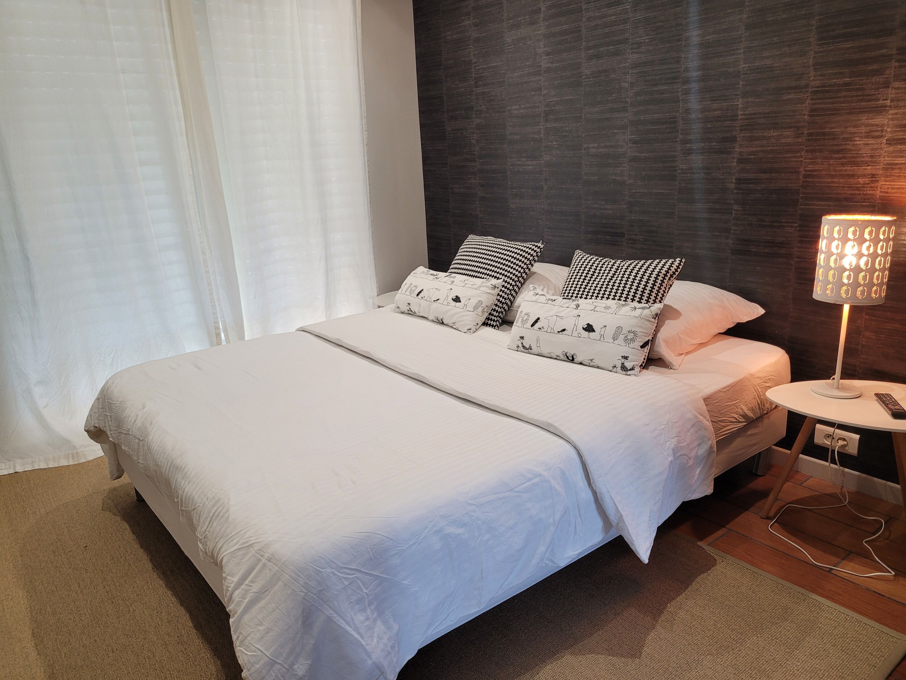 A neatly made bed with white bedding and patterned pillows, a bedside table with a lamp, and a dark wooden headboard.
