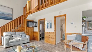 A spacious living area with a wooden staircase, a grey sofa, a glass coffee table, and a wooden chair. - Serenity Now: Oceanside | Screened in Porch | (Duck)