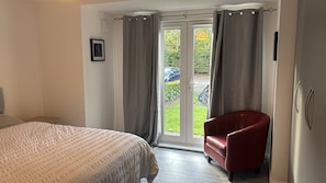 A bedroom with a bed, a red chair, and a sliding glass door leading to a garden. - Quiet Ground-Floor Apartment | Heathrow, Windsor & London at Your Doorstep (Slough)