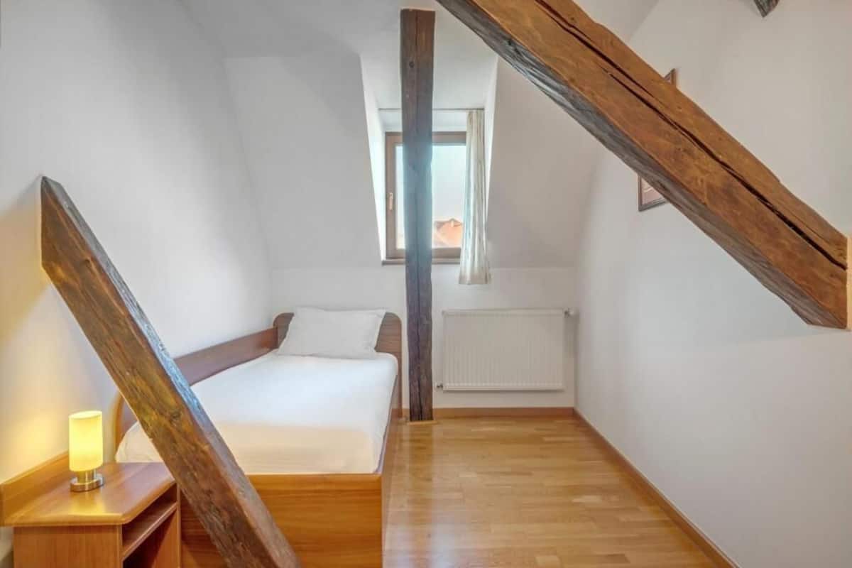 A bedroom with a wooden ceiling beam, a bed, a nightstand with a lamp, and a window with a view of a building outside.