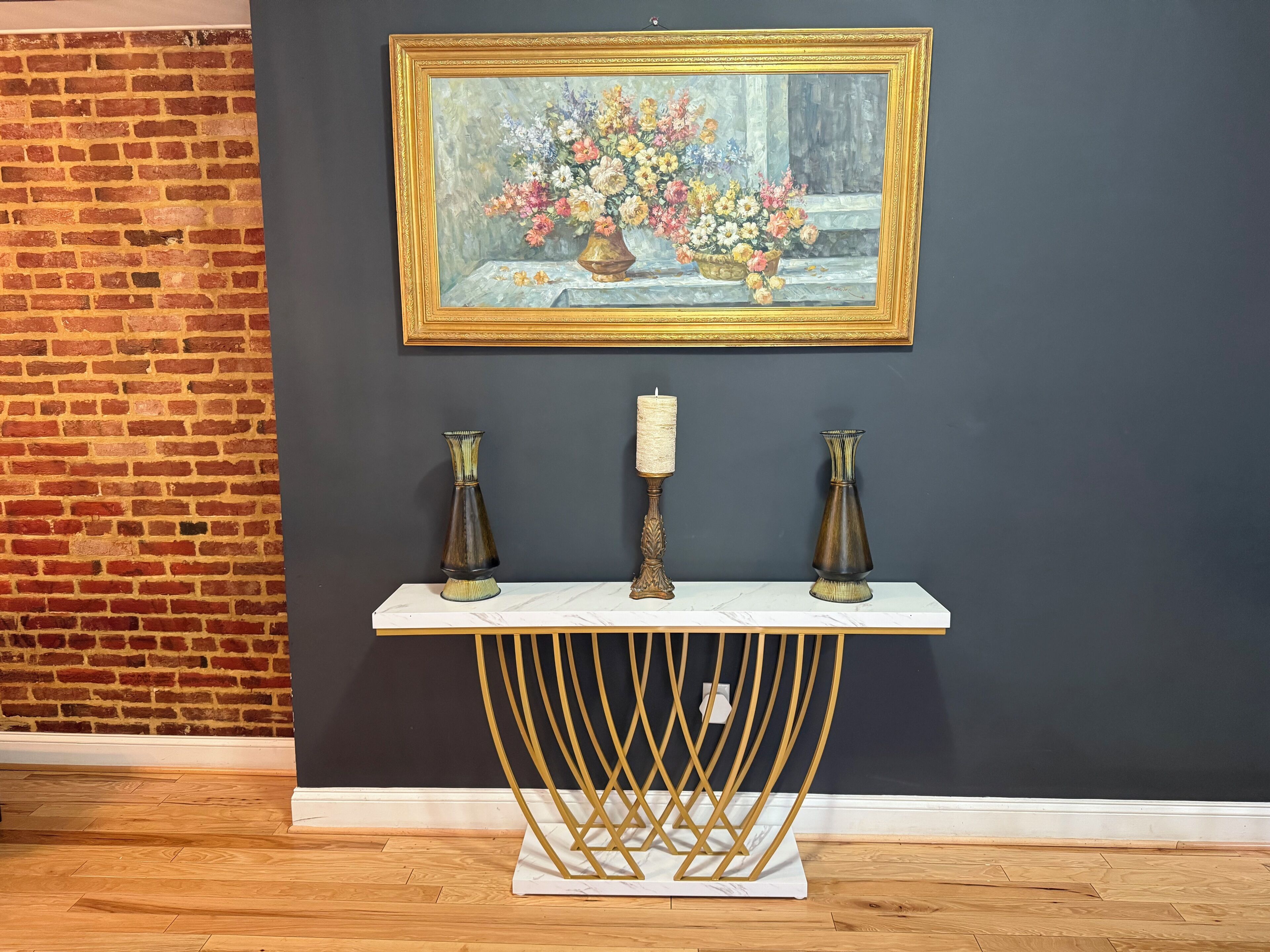 Quarto com parede de tijolos, um quadro de flores, um console branco com vasos e uma vela.