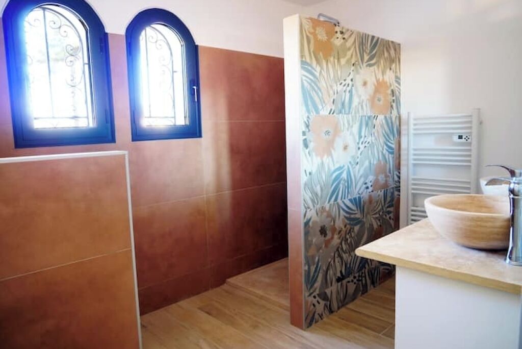 Ein Badezimmer mit zwei Rundbogenfenstern, einer Dusche mit floralem Fliesenmuster und einem hölzernen Waschbecken.