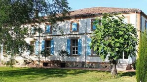 Un bâtiment à deux  étages, avec un toit recouvert de tuiles et des murs blancs. Les fenêtres sont dotées de volets bleus, et un arbre se trouve au premier plan.