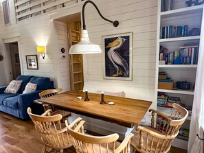 A dining area with a wooden table, wicker chairs, a white pendant light, a bookshelf, and a framed pelican painting. - CR61: Cedar Tree Cottage: Historic Charm | Wheelchair Ramp (Ocracoke)