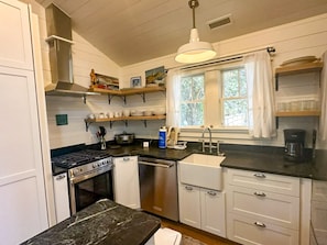 A kitchen with white cabinets, a black countertop, a stove, a sink, and a window with curtains. - CR61: Cedar Tree Cottage: Historic Charm | Wheelchair Ramp (Ocracoke)