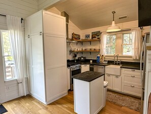 A compact kitchen with white cabinets, a black countertop, and a window with curtains. - CR61: Cedar Tree Cottage: Historic Charm | Wheelchair Ramp (Ocracoke)
