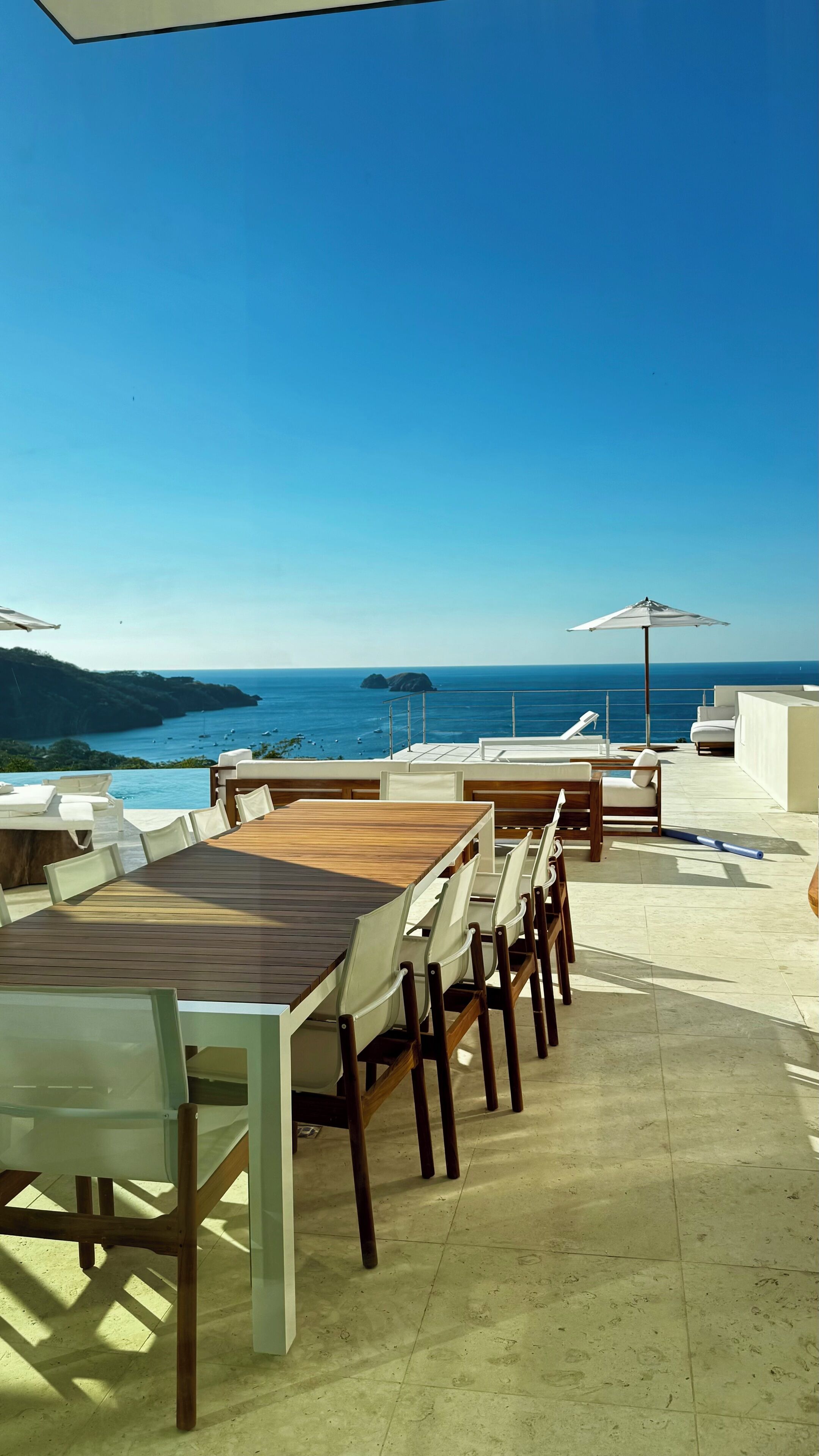 Une terrasse extérieure avec une table et des chaises en bois, un parasol blanc et une vue sur la mer.