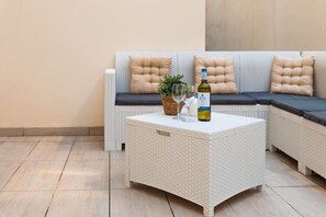 A wicker sofa with cushions, a small table with a bottle of wine, a glass, and a plant. - Ortigia Collection / Suite No.1 / Terrace (Swieqi)