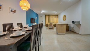 A modern living room with a dining area, a TV mounted on the wall, a kitchen area with a microwave, and a blue accent wall. - Ortigia Collection / Suite No.1 / Terrace (Swieqi)