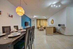 Een moderne woonkamer met een eethoek, een televisie aan de muur en een keuken met een magnetron, plus een blauwe accentmuur.
