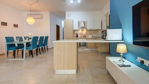 A modern kitchen with a central island, a dining area with blue chairs, and a blue accent wall. - Ortigia Collection / Suite No.5 / Terrace (Swieqi)