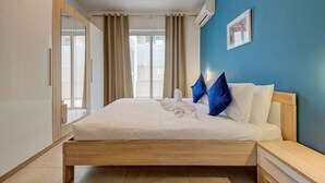 A hotel room with a bed, a bedside table, a mirror, a window with curtains, and an air conditioning unit. - Ortigia Collection / Suite No.4 / Terrace (Swieqi)