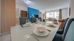 A modern dining area with a glass table, blue chairs, and a blue accent wall. - Ortigia Collection / Suite No.4 / Terrace (Swieqi)