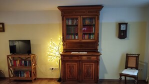 Uma sala de estar com uma estante de madeira, uma televisão sobre um suporte, uma luminária decorativa, um relógio na parede e uma cadeira.