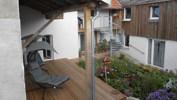 Um deck de madeira com uma cadeira, um pequeno jardim com flores e uma escada que leva ao segundo andar.